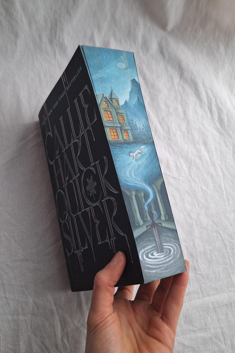 May include: A hardcover book titled "Quicksilver" by Callie Hart. The cover features a detailed illustration of a snowy landscape with a house, a wolf, and a sword. The title is embossed in silver on a black background.