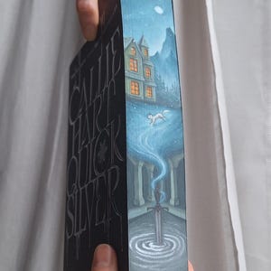 May include: A book with a dark cover featuring the title "Callie Hart Quicksilver" in silver lettering. The spine has a painted illustration of a house, a running fox, and a sword in a pool of water, under a starry night sky.