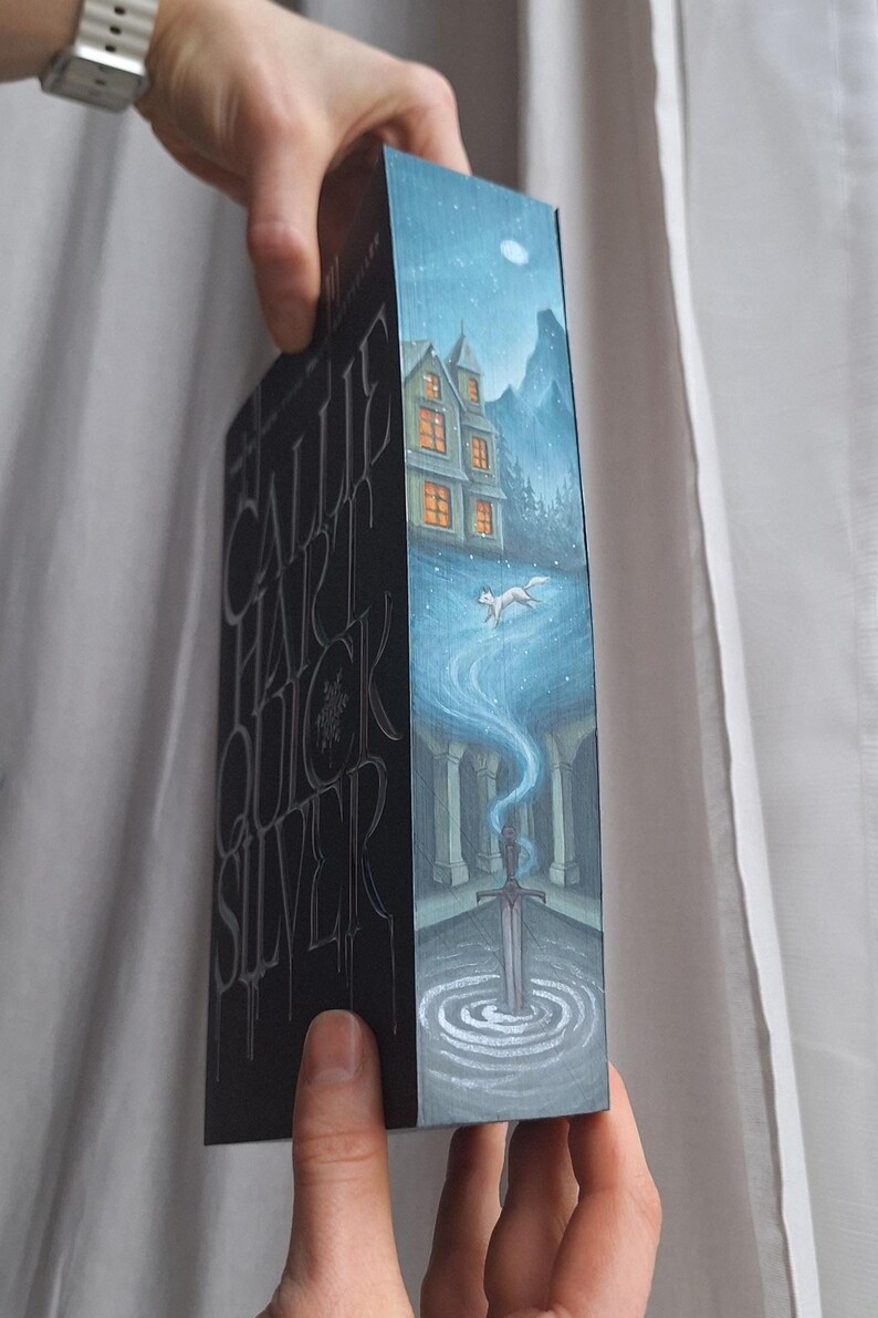 May include: A book with a dark cover featuring the title "Callie Hart Quicksilver" in silver lettering. The spine has a painted illustration of a house, a running fox, and a sword in a pool of water, under a starry night sky.