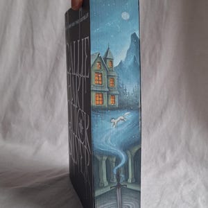 May include: A book spine with a detailed illustration. The artwork depicts a house with lit windows, a running fox, and a sword in a pool of water. The background is a night scene with a moon and stars. The book title is visible.
