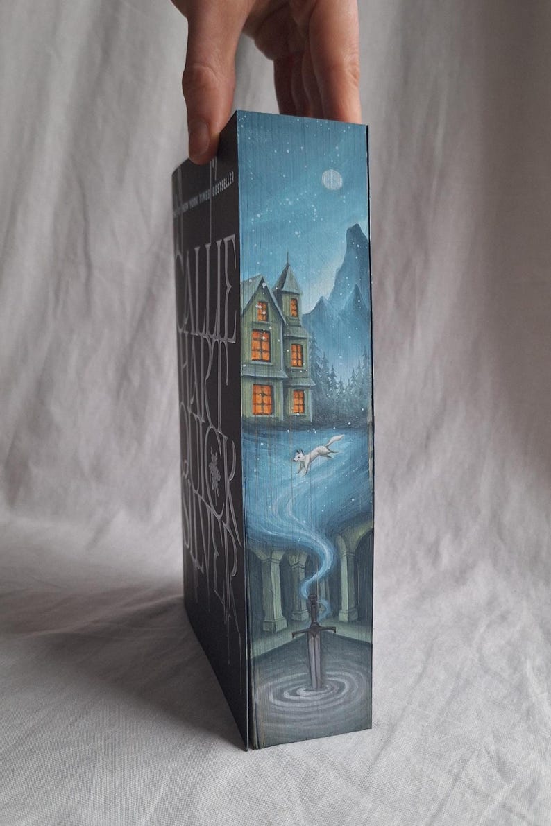 May include: A book spine with a detailed illustration. The artwork depicts a house with lit windows, a running fox, and a sword in a pool of water. The background is a night scene with a moon and stars. The book title is visible.