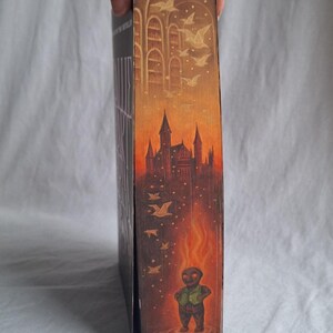 May include: A book with a detailed illustration on the side. The artwork depicts a castle, birds, and a figure engulfed in flames. The color palette includes shades of orange, brown, and gold, creating a fantasy theme.