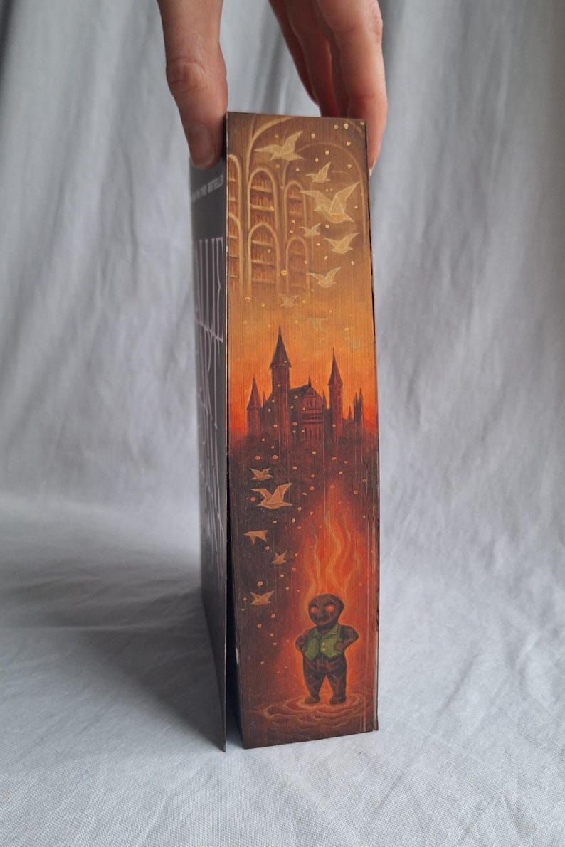 May include: A book with a detailed illustration on the side. The artwork depicts a castle, birds, and a figure engulfed in flames. The color palette includes shades of orange, brown, and gold, creating a fantasy theme.