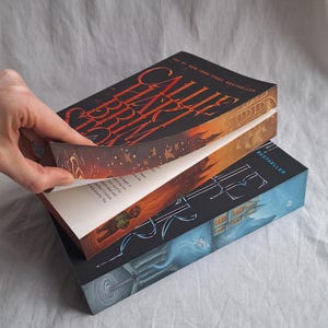 May include: Three hardcover books are stacked, with the top book's pages being turned. The top book has a black cover with the title "Callie Hart Brim" in red. The second book has a blue cover with a building illustration.