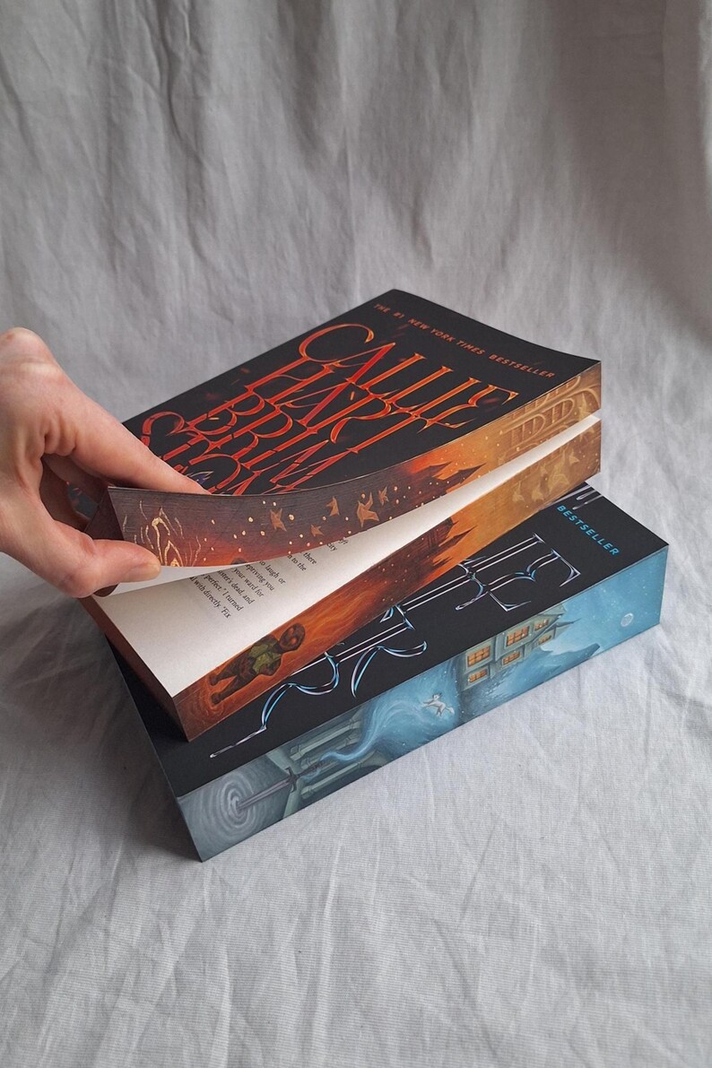May include: Three hardcover books are stacked, with the top book's pages being turned. The top book has a black cover with the title "Callie Hart Brim" in red. The second book has a blue cover with a building illustration.