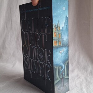 May include: A hardcover book titled "Quicksilver" by Callie Hart. The cover is black with silver lettering and a wintery illustration of a house, mountains, and a fox. The text "#1 New York Times Bestseller" is at the top.
