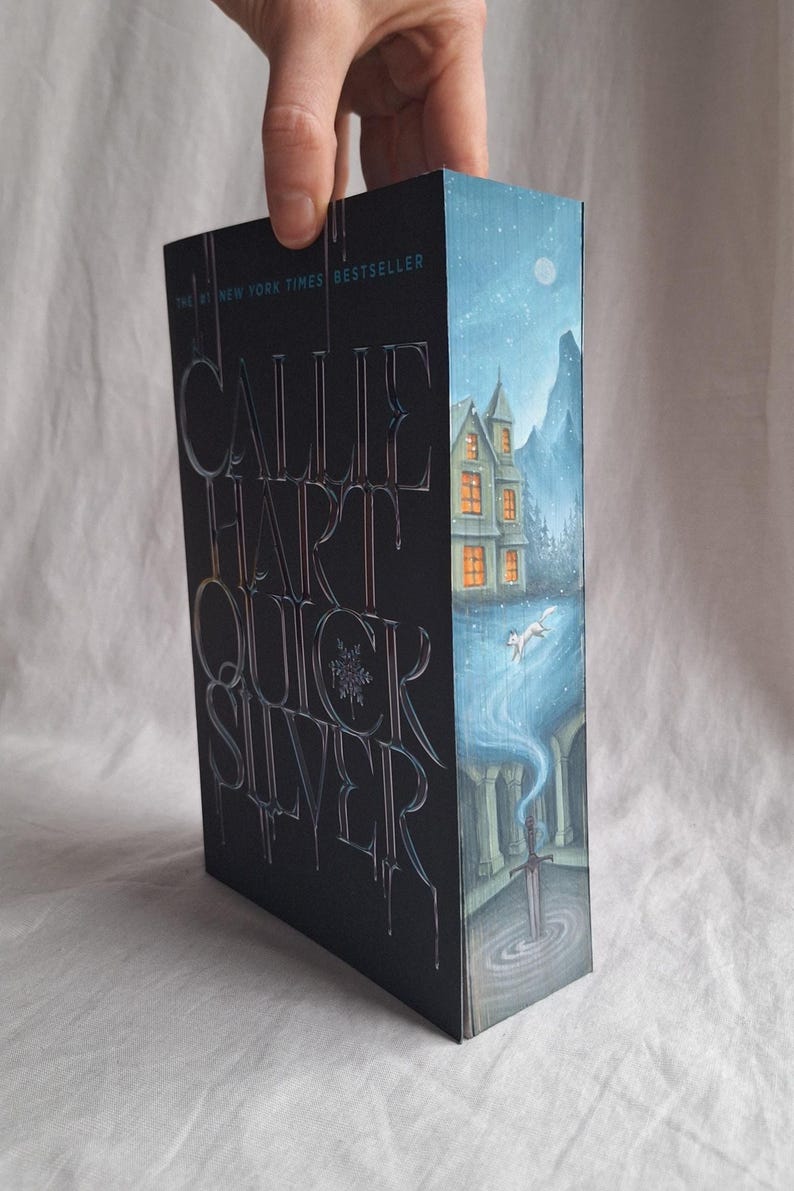 May include: A hardcover book titled "Quicksilver" by Callie Hart. The cover is black with silver lettering and a wintery illustration of a house, mountains, and a fox. The text "#1 New York Times Bestseller" is at the top.