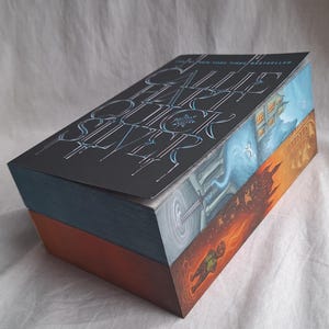 May include: A hardcover book with a dark cover featuring the title "CALL ME BY YOUR NAME" in stylized, light blue lettering. The book's edges are painted with colorful scenes, including a house, a portal, and a figure in flames.