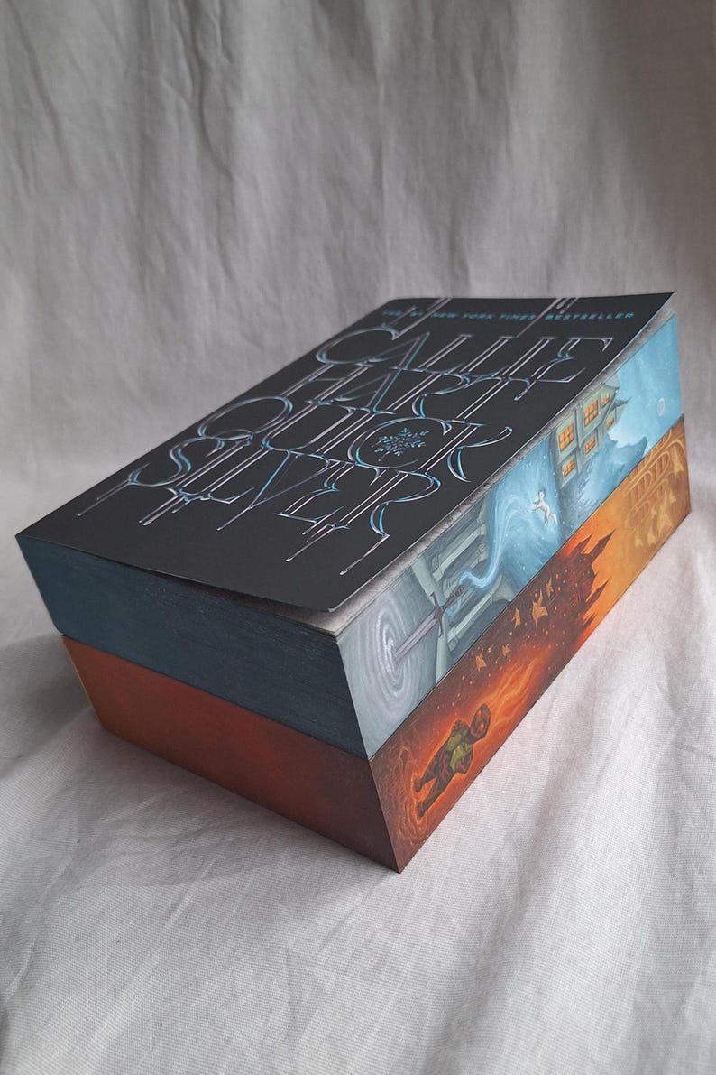 May include: A hardcover book with a dark cover featuring the title "CALL ME BY YOUR NAME" in stylized, light blue lettering. The book's edges are painted with colorful scenes, including a house, a portal, and a figure in flames.