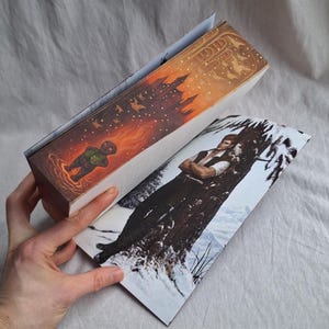 May include: A book with a colorful illustration on the cover and spine. The spine features a castle silhouette against an orange sky. The cover shows a man leaning against a tree in a snowy landscape. The book is held open by a hand.