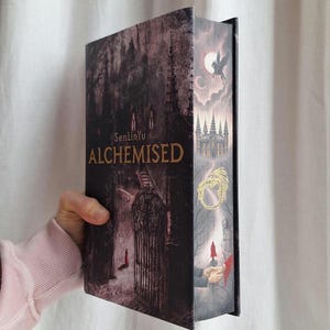 May include: Hardback book titled "ALCHEMISED" by SenLinYu. The cover features a dark, gothic illustration with a castle, a winged creature, and a figure in a long coat. The colour palette is primarily dark red, black, and gold.