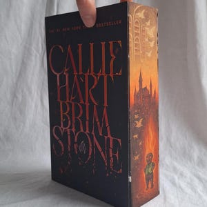 May include: A hardcover book titled "CALLIE HART BRIMSTONE" with a dark cover and red lettering. The spine features a castle illustration in shades of orange and yellow. The book is a New York Times bestseller.