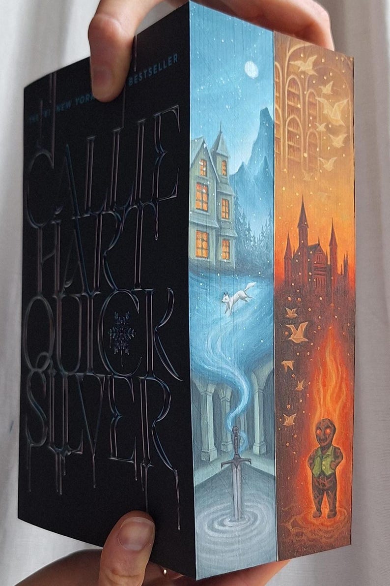 May include: A hardcover book titled "Quicksilver" by Callie Hart. The book's spine is black with silver lettering. The edges of the pages are painted with fantasy scenes in blue, orange, and brown tones, featuring a house, a fox, a sword, and a figure.