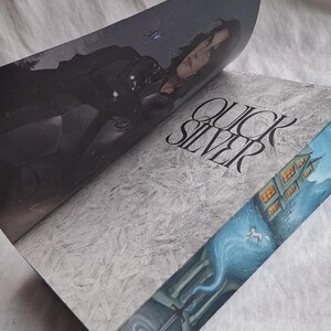 May include: Open book with the title "QUICKSILVER" in a stylized font. The cover features a woman in dark clothing, while the edges showcase a detailed illustration of a building and a swirling blue form.