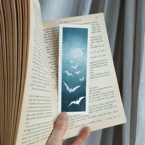以下が含まれることがあります： 満月を背景にコウモリが飛んでいる青と白のイラストが描かれたしおりが付いた開いた本のクローズアップ。しおりには「メリークリスマス」の文字が書かれている。
