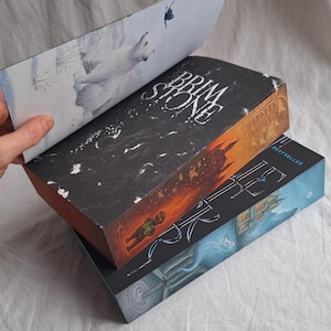 May include: Three hardcover books stacked, with the top book partially open. The top book's cover is black with white text, and the open page shows a polar bear illustration. The middle book has an orange and black cover, and the bottom book is blue.