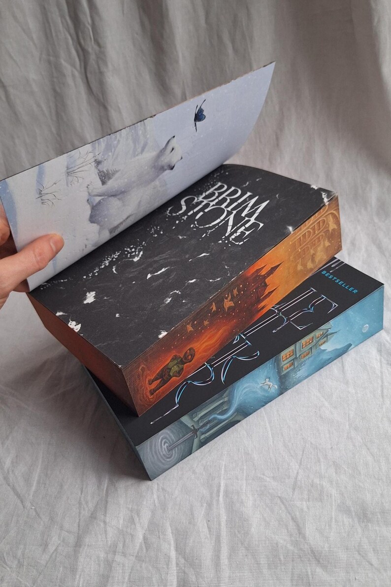 May include: Three hardcover books stacked, with the top book partially open. The top book's cover is black with white text, and the open page shows a polar bear illustration. The middle book has an orange and black cover, and the bottom book is blue.