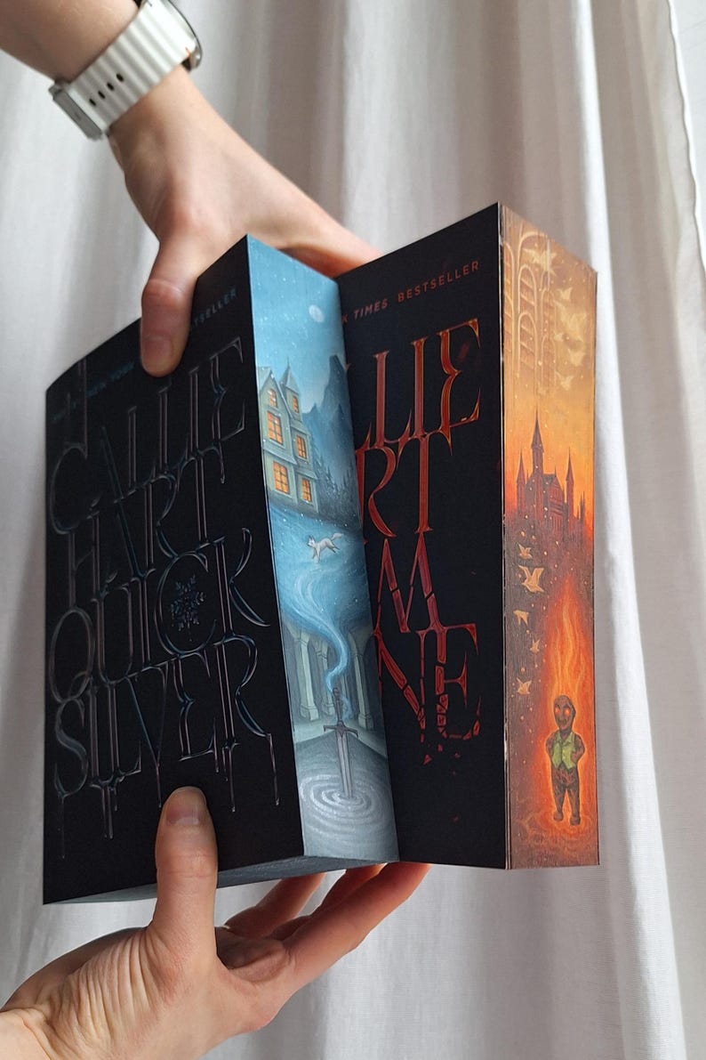 May include: A person holding open a book with a black cover and embossed silver lettering. The book's pages reveal colorful illustrations of a snowy landscape, a sword, and a burning castle. The book's title is "Callie Hart Quicksilver".