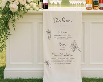 Peut inclure: Un bar blanc avec une bannière crème indiquant "The bar". La bannière liste les options de vin, de bière et de boissons non alcoolisées. Des bouteilles de vin, des verres et des fleurs sont visibles.