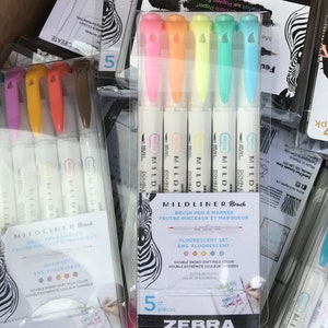 Puede incluir: Un paquete de cinco bolígrafos de pincel Zebra Mildliner en colores fluorescentes. Los bolígrafos tienen una punta doble y son resistentes al agua. El embalaje presenta un diseño de estampado de cebra y el texto "Mildliner Brush" y "Fluorescent Set".