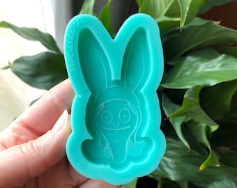 May include: A teal blue silicone mold with a raised design of a cartoon bunny wearing a headband. The bunny has large eyes and a small mouth.