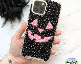 May include: A clear phone case with a black and pink pumpkin design made of rhinestones. The case is decorated with black and pink gems.