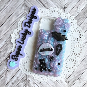 Puede incluir: Una funda transparente para teléfono con fondo morado y azul. La funda está decorada con abalorios de temática de Halloween en blanco y negro, incluyendo un murciélago, un ataúd, una araña, un osito de goma y una boca de vampiro.