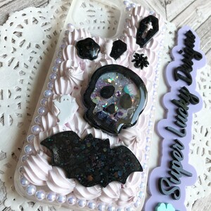 Puede incluir: Una funda de teléfono blanca decorada con adornos de Halloween en blanco y negro, incluyendo una calavera, un murciélago, un fantasma y un ataúd. La funda también está decorada con glaseado blanco y cuentas iridiscentes. El texto "Super Lucky Designs" está escrito en negro sobre un fondo morado.