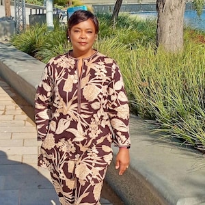 May include: A woman wearing a brown and beige floral print pantsuit with a long-sleeved top and matching pants. She is walking on a paved path with a stone wall and green plants in the background.