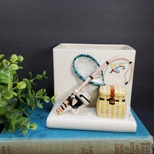 Puede incluir: Maceta de cerámica blanca con temática de pesca. La maceta presenta una caña de pescar, una red y una caja de aparejos en relieve. La maceta está sentada sobre un libro azul.