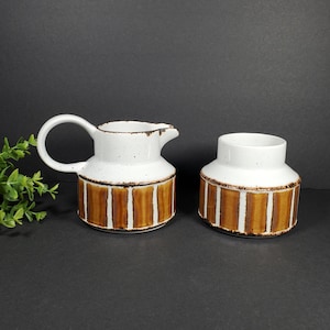 May include: A white ceramic creamer and sugar bowl set with brown and white striped designs. The creamer has a handle and a spout. The sugar bowl is round.