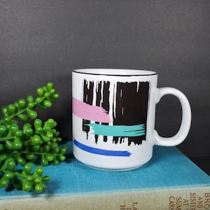 May include: White ceramic mug with a black rim and abstract design featuring black, pink, blue, and teal brushstrokes.