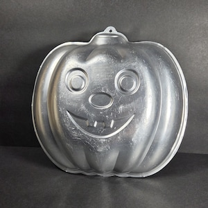 May include: A silver-colored metal cake pan shaped like a jack-o'-lantern pumpkin. The pan features a molded face with two circular eyes, an oval nose, and a wide, smiling mouth with two square teeth. It has a small hole at the top for hanging.
