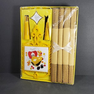 May include: A vintage yellow boxed set featuring a decorative tile with fruit illustrations, yellow serving utensils, and woven placemats. The placemats are tied with a white ribbon. The set is in its original packaging.