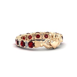 May include: A gold Claddagh ring featuring a central heart topped with a crown and held by two hands. The band is set with alternating round red rubies and small clear white diamonds, creating a continuous circular pattern around the finger.
