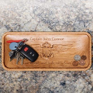 May include: A rectangular wooden tray with a rope-like border. The tray is engraved with the words "Captain John Connor" and "Fair Winds and Following Seas". A set of keys and coins rest on the tray.
