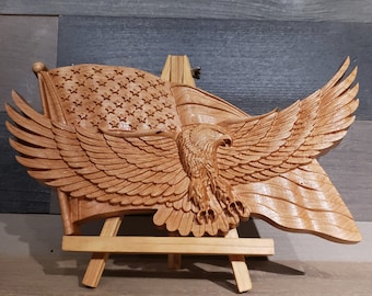May include: A wooden carving of an American bald eagle with its wings spread wide, perched atop a wooden American flag. The carving is displayed on a small wooden easel.