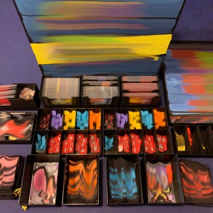 May include: A collection of black plastic storage boxes with colorful card decks, dice, and tokens. The boxes are organized on a blue surface.