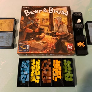 May include: A board game called "Beer & Bread" with a box featuring a medieval-themed illustration of two people drinking beer and bread. The game includes a variety of wooden pieces in blue, yellow, brown, and green.
