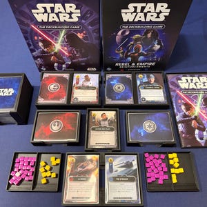 Könnte beinhalten: Ein Star Wars The Deckbuilding Game Set mit zwei Spielschachteln, einem Regelbuch, Kartenstapeln und Kunststoffschalen mit kleinen rosa und gelben quadratischen Spielsteinen. Die Spielkomponenten zeigen Charakterbilder und Text.