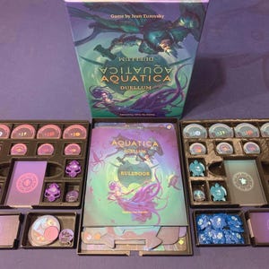 以下が含まれることがあります： ボードゲーム「Aquatica Duellum」の箱とコンポーネント。箱には水棲生物が描かれています。内部には、紫、青、ピンク色のゲームピース、カード、トレイがあります。ゲームはプレイできる状態のようです。