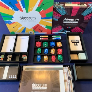 May include: A game box set for the board game "décorum: Movin' Out". The box set includes a game board, cards, tokens, and a rulebook. The game is an expansion for the original "décorum" game.