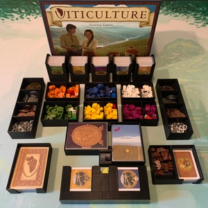 May include: A black and brown board game organiser with compartments for storing game pieces and cards. The organiser is labelled "Viticulture: Essential Edition".