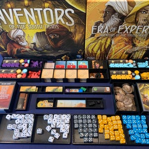 May include: Board game titled "Inventors of the South Tigris" and "Era of Experts." The game includes dice, cards, tokens, and game pieces in black trays. The game boxes feature illustrations of figures in Middle Eastern attire.