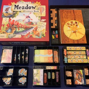 以下が含まれることがあります： 「Meadow Adventure Book」と書いてある赤い箱。箱の中には、ルールブック、カード、トークンなど、さまざまなコンポーネントが含まれたボードゲームが入っています。このゲームは1〜4人でプレイでき、10歳以上のお子様向けです。