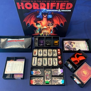 May include: The board game "Horrified: Dungeons & Dragons" is displayed with its components. The box art features a dragon and the text "Terror is in the Eye of the Beholder!". The game's contents are organized in black trays, including cards, figures, and dice.