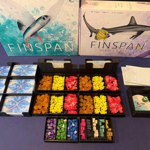 Könnte beinhalten: Zwei Brettspielboxen, "Finspan" und "Finspan Sharks & Reefs", mit Spielkomponenten. Die "Finspan"-Box zeigt einen Fisch mit Flügeln, während die "Sharks & Reefs"-Box einen Hai zeigt. Zu den Komponenten gehören Karten, Spielsteine und Spielteile.
