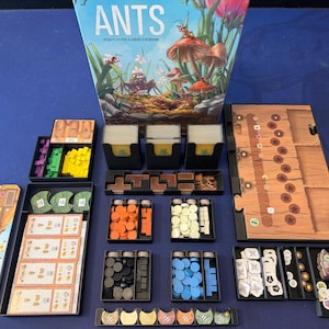 May include: The board game ANTS by Renato Ciervo and Andrea Robbiani, featuring a box with an illustration of ants and mushrooms, includes various black plastic trays containing orange, yellow, green, blue, black, and white tokens and cards.