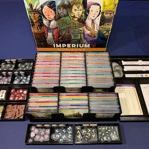May include: A black plastic organizer with multiple compartments for storing game pieces and cards. The organizer is filled with various game components, including cards, tokens, and dice. The box for the game "Imperium: Horizons" is visible in the background.