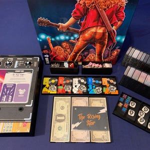 May include: Board game with a rock and roll theme. The game includes a board, cards, money, and various game pieces. The box art features a musician playing guitar in front of a crowd. The game components are arranged on a blue surface.